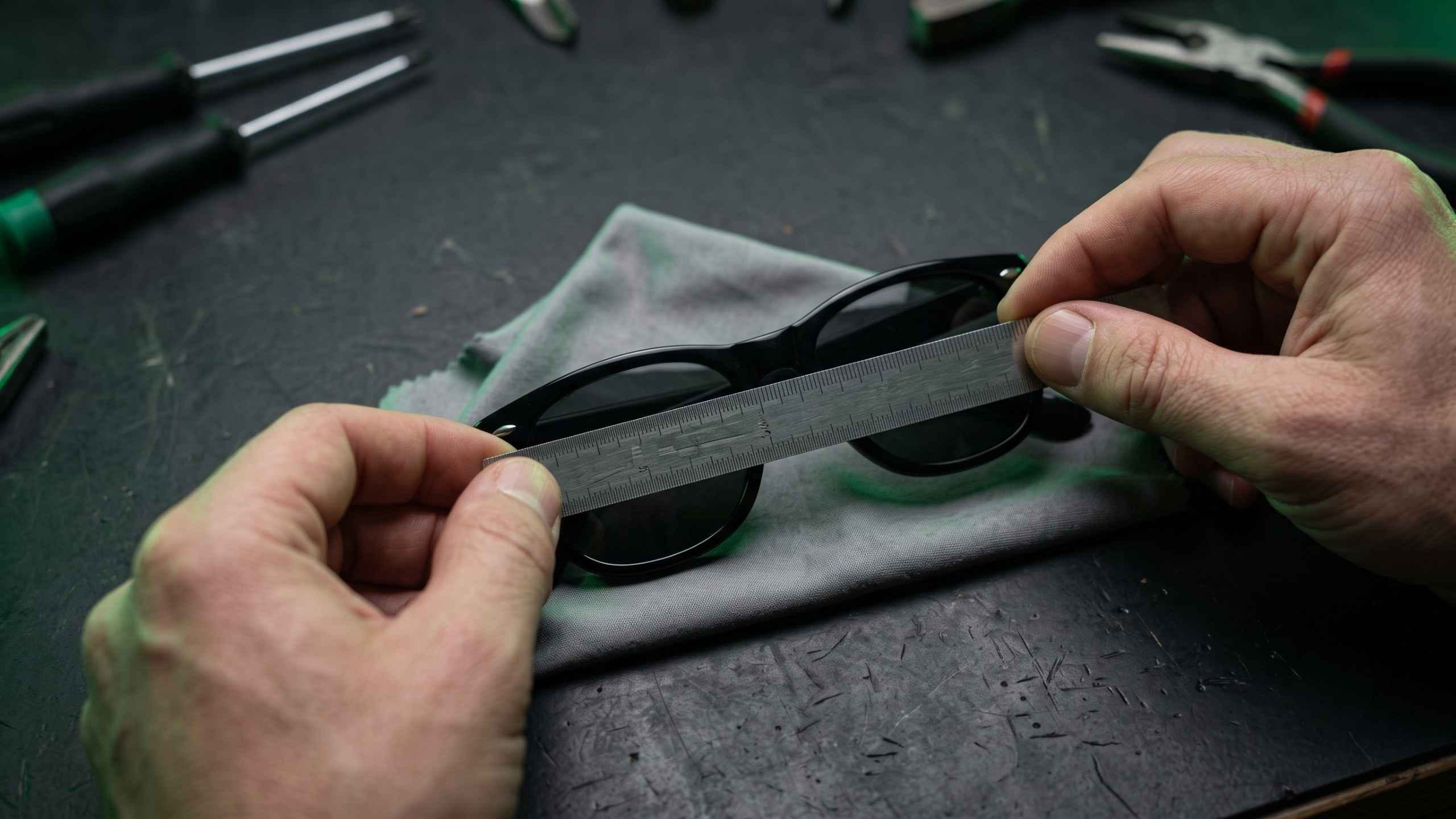 Hands measuring the width of a sunglass lens with a steel ruler on a dark workbench